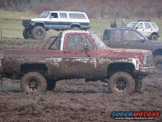 Muddy red GMC pickup truck with roll bar, parked next to a lifted white SUV and a dark pickup truck in a muddy field