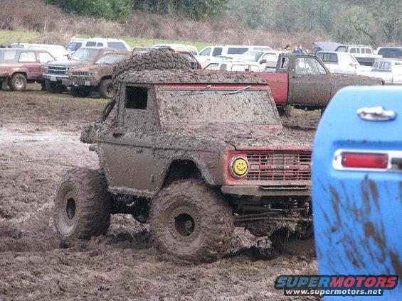 Muddy 1978 Ford Bronco with large tires in a mud pit, other vehicles in background