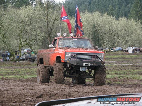 Orange lifted pickup truck with large tires in mud, flying two Confederate flags