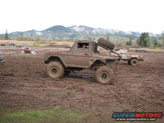 Muddy off-road vehicle with spare tire on back, driving through a muddy field with other vehicles