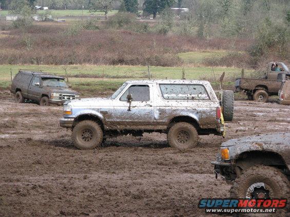 White SUV stuck in mud, side view. Text on rear window reads "SuperMotors.net"