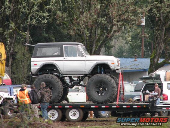 Lifted 1978 Ford Bronco with oversized tires on a flatbed trailer, side view. Other vehicles and trees in background