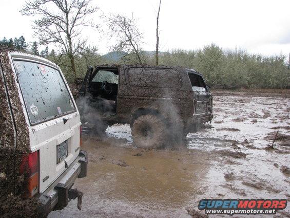 Two muddy SUVs stuck in a muddy field. One is a dark-colored SUV with its door open, the other is a white SUV seen from the rear