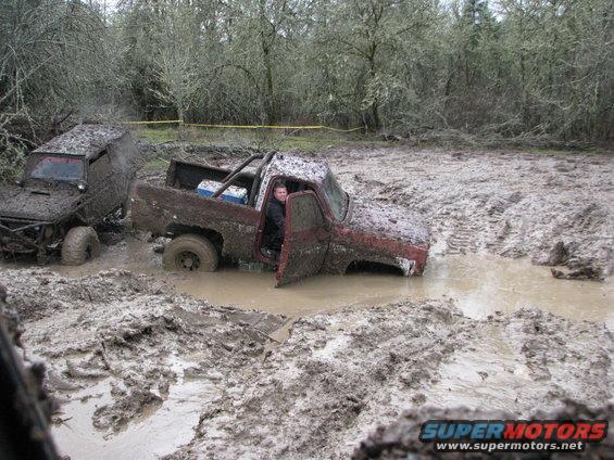 Muddy red pickup truck with open door stuck in mud next to another muddy vehicle