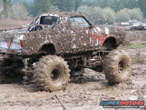 Mud-covered monster truck with huge tires stuck in mud. Some red paint visible on the side