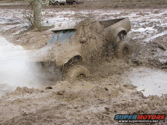 Truck deep in mud, tires spinning, throwing up spray. Another vehicle visible in background