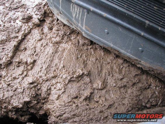 Close-up of a vehicle tire deeply embedded in thick mud. The lower body panel of the vehicle is visible above the mud