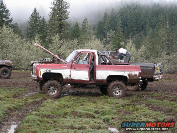 Red and white pickup truck with hood up, people working on it in a muddy field