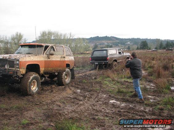 Mud-covered orange and white 1978 Ford Bronco with large tires, and a black 1978 Ford Bronco being pulled by a rope in a muddy field