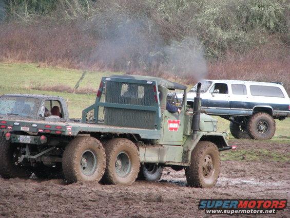 Military truck with six wheels towing a dark SUV through mud, with a lifted white SUV in the background