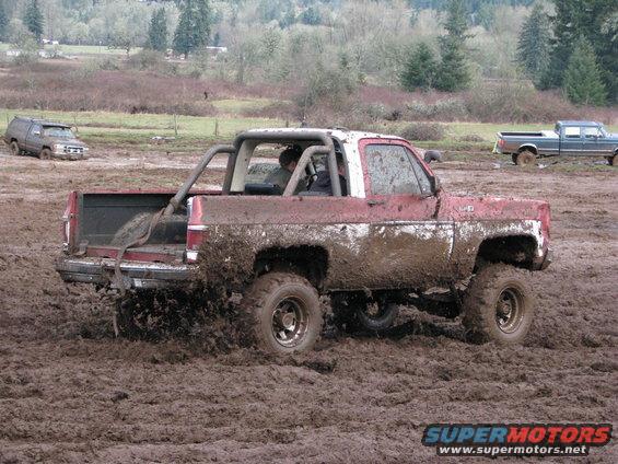 red pickup truck with roll cage driving through mud, another SUV and truck in background