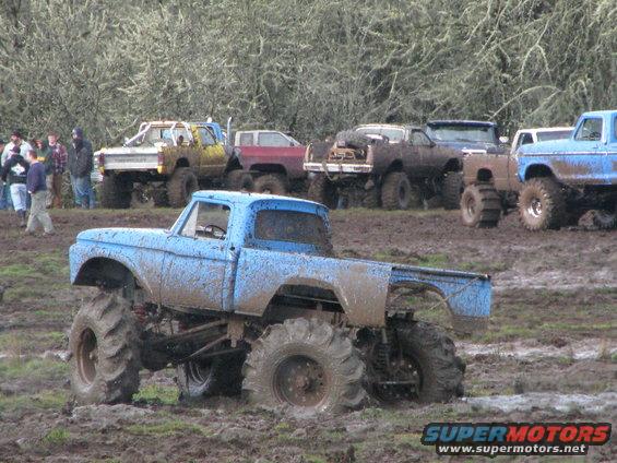 Mud-covered blue pickup truck with oversized tires stuck in mud