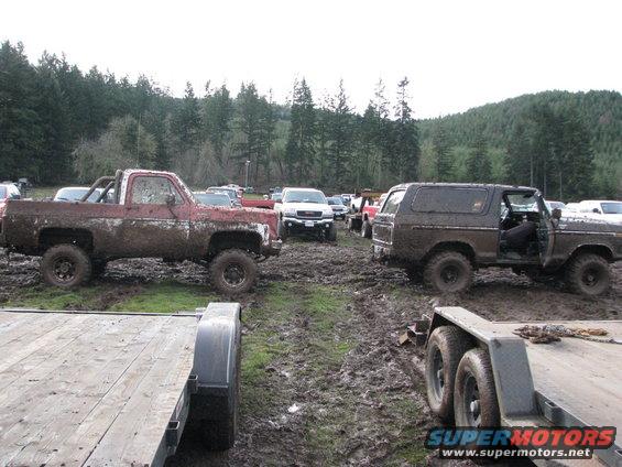 Muddy 1978 Ford Bronco parked next to a red pickup truck in a muddy field with trees in the background