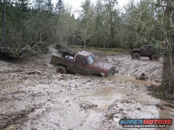Red pickup truck stuck deep in mud, another off-road vehicle in background