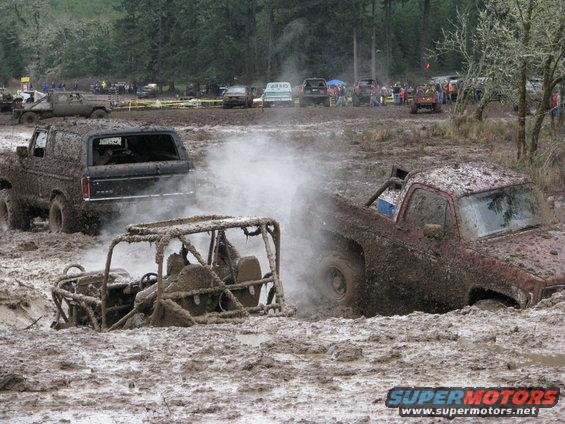 Mud-covered 1978 Ford Bronco with smoke coming from behind, and a red pickup truck stuck in mud