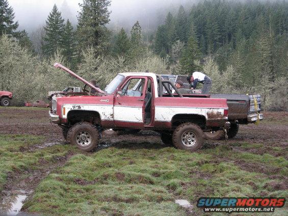 Red and white pickup truck with hood open, stuck in mud. Another truck is behind it. Trees and fog in background