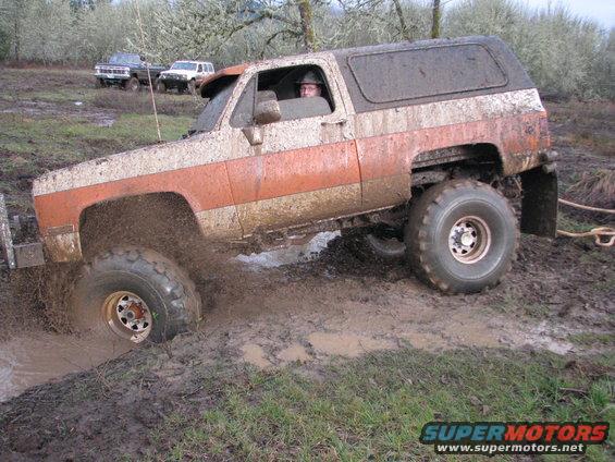 Mud-splattered off-road SUV with orange and white paint stuck in mud, other vehicles in background