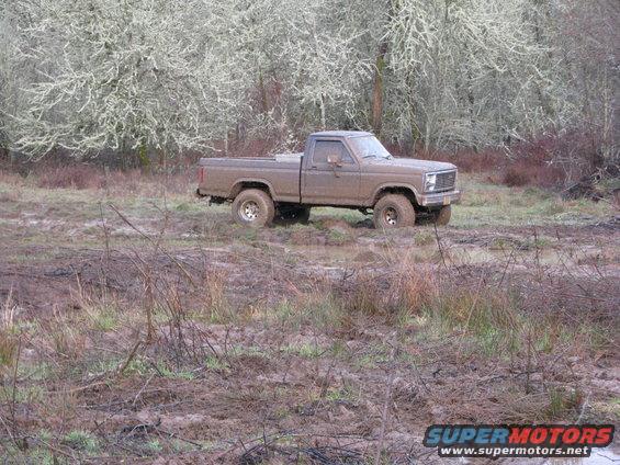 Brown pickup truck stuck in mud, front quarter view. Muddy tires and body. Bare trees in background