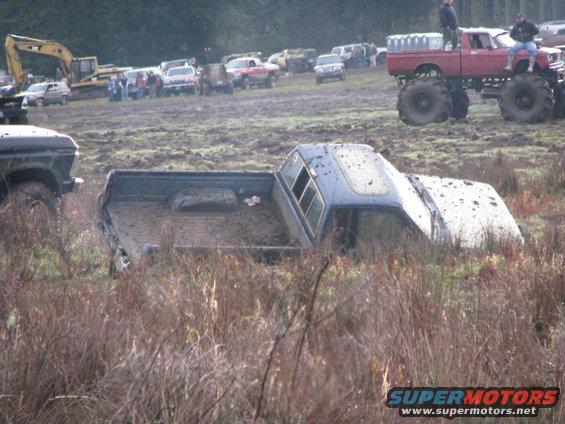 Blue pickup truck stuck in mud, side view. Other vehicles and excavator in background