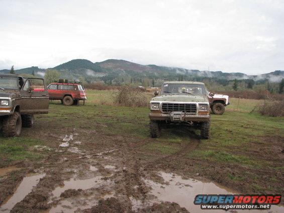 Green Ford F-series truck with winch in front, brown Ford truck to left, red Jeep Wagoneer in background, muddy field