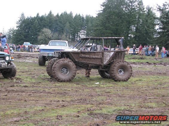Muddy custom buggy with large tires, parked in a muddy field next to a pickup truck and other vehicles