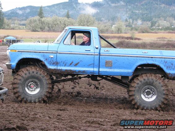 Blue pickup truck with large tires in mud. A man is driving. The truck has a roll bar