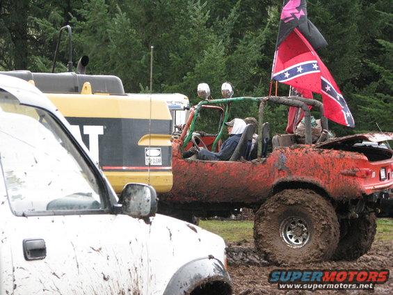 Muddy off-road buggy with Confederate flag, next to a white pickup and yellow excavator