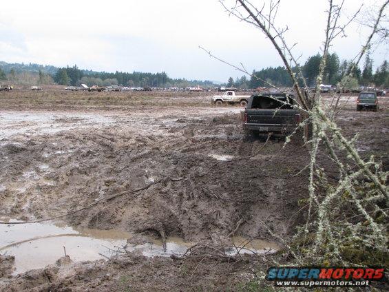 Black pickup truck stuck in deep mud, rear view. Another white truck is visible in the background