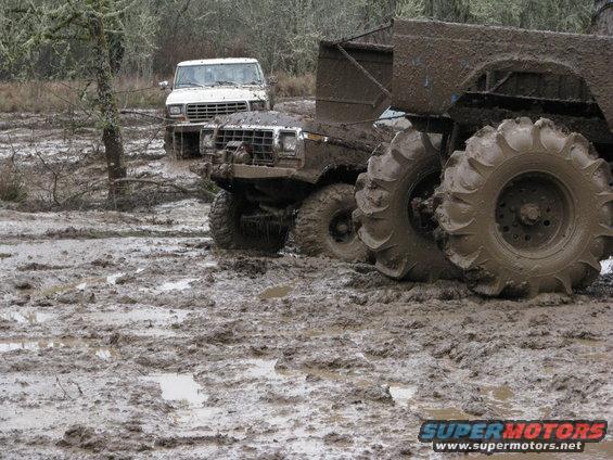 Two trucks stuck in mud, one in foreground with large tires, another white truck behind it