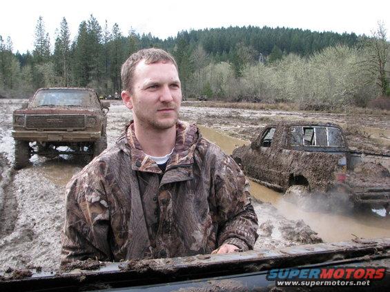 Man in camouflage jacket with muddy trucks stuck in mud; one truck is a dark pickup, the other is a lighter-colored pickup