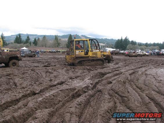 John Deere bulldozer stuck in mud, with other vehicles and trees in background