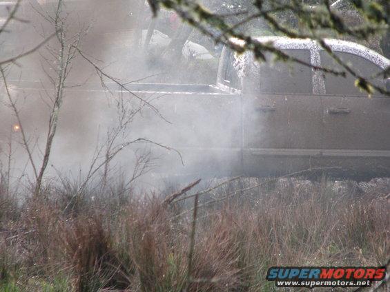 Mud-covered pickup truck side view, partially obscured by smoke and vegetation