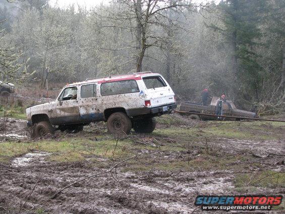 White SUV stuck in mud, being towed by brown pickup truck with two people nearby