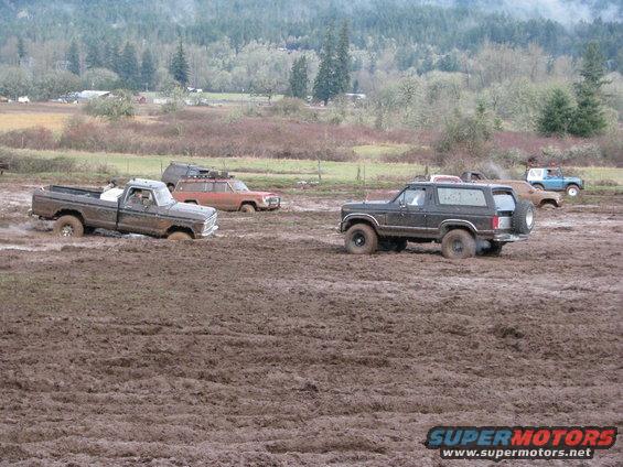 Several off-road vehicles stuck in thick mud, with trees and hills in the background