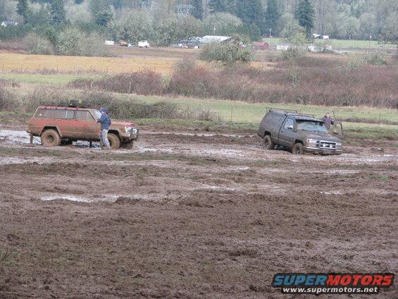 Two SUVs are stuck in a muddy field, one red and one dark gray