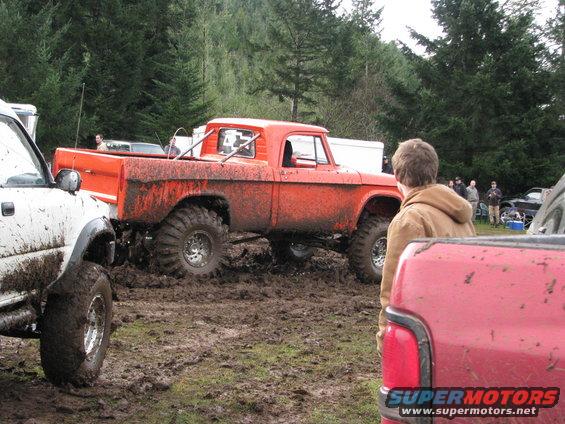 Orange pickup truck with large tires stuck in mud, white truck to the left, person watching