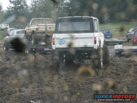 White Ford Bronco stuck in mud, towing a trailer with a pickup truck