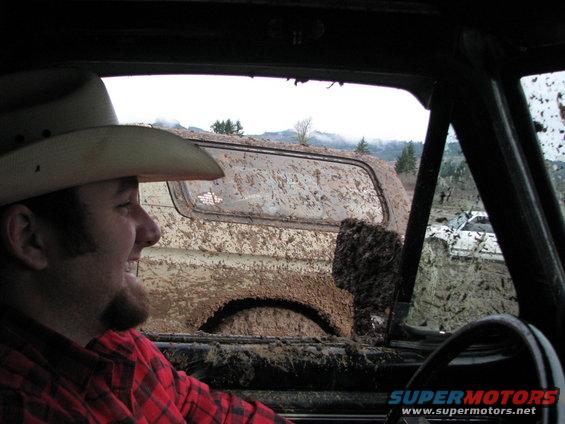 Man in cowboy hat looks out window at muddy off-road vehicles, one is white, in a muddy field
