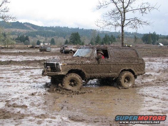 Mud-covered 1978 Ford Bronco driving through a muddy field with other vehicles in the background