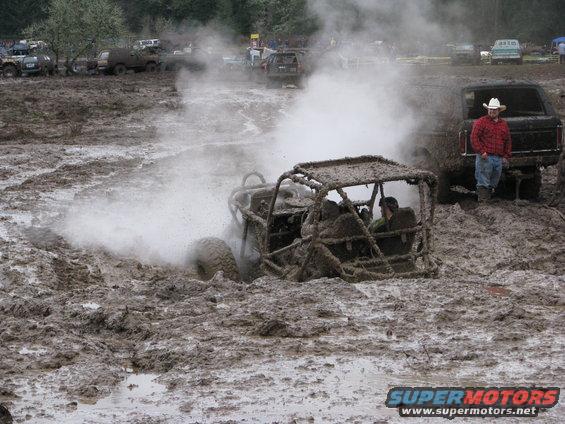 Mud-covered buggy spinning wheels in thick mud, with a man standing by a black SUV in the background
