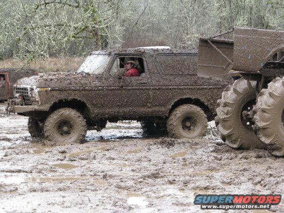 Mud-covered 1978 Ford Bronco driving through a muddy field with another vehicle partially visible