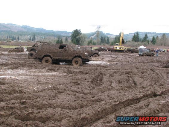 Muddy off-road vehicles stuck in a muddy field with a yellow excavator in the background