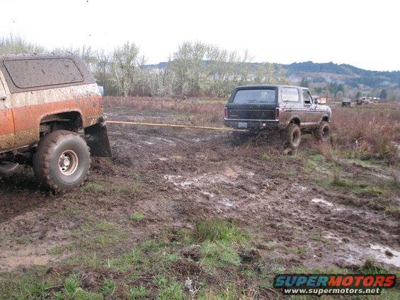 Black 1978 Ford Bronco being towed by an orange and white off-road vehicle in a muddy field