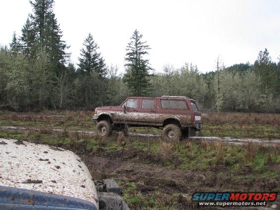 Mud-covered maroon Ford Bronco with a camper shell driving through a muddy field