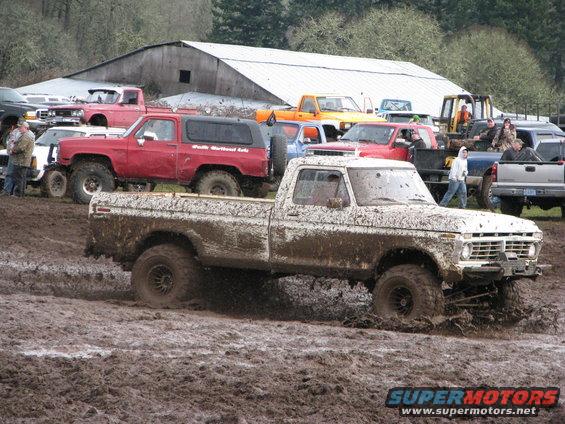 Muddy 1977 Ford F-Series pickup truck with lifted suspension driving through deep mud. Other trucks and a barn are in the background