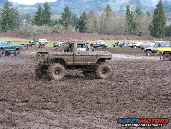 Mud-covered truck with huge tires in a muddy field, other vehicles and trees in background