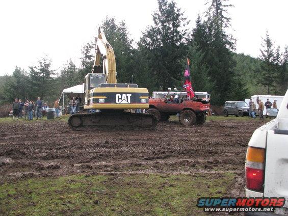 Excavator and red off-road vehicle in muddy field with trees and people
