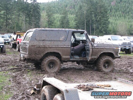 Muddy 1978 Ford Bronco with door open, parked in a muddy field with other vehicles around