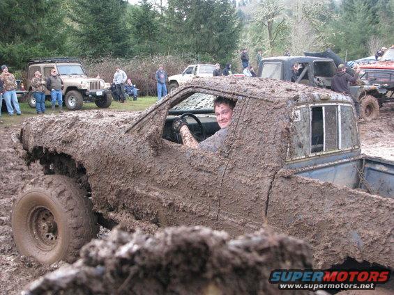 Mud-covered pickup truck with driver, other off-road vehicles in background