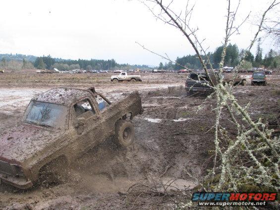 Mud-covered pickup truck struggling in a mud pit, other vehicles visible in background